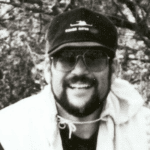A smiling man wearing sunglasses, a cap, and casual outdoor clothing outdoors in front of trees, representing citizen journalism and free speech advocacy.