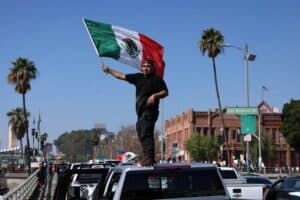 Man holding Mexican flag atop vehicle during parade in Los Angeles, demonstrating patriotism and cultural pride.